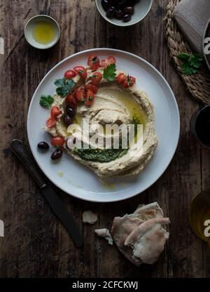 Da sopra piatto di delizioso hummus pesto decorato con ciliegia pomodori e fagioli con prezzemolo messo su tavola di legno vicino pane piatto e olio Foto Stock