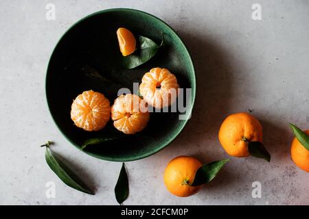 Vista dall'alto della ciotola in ceramica verde con mandarini sbucciati freschi posto su tavola bianca vicino a frutta non sbucciata con foglie verdi Foto Stock