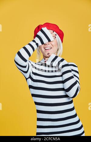 Allegro bionda Donna in blusa a righe e cappellino rosso francese sorridente con le mani sul viso su sfondo giallo Foto Stock
