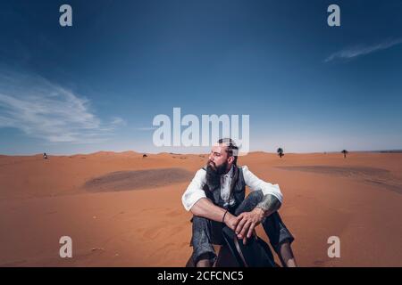 maschio che tiene il cappello del cowboy e che siede sulla sabbia contro il deserto e cielo blu Foto Stock