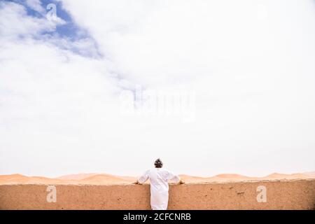 Vista posteriore di anonimo uomo adulto in lunghi abiti bianchi poggiati su un muro che guarda lontano contro l'infinito deserto di sabbia, il Marocco Foto Stock