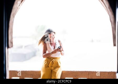 Donna felice usando il telefono cellulare vicino al paesaggio desertico in piedi sul balcone di pietra, Marocco Foto Stock