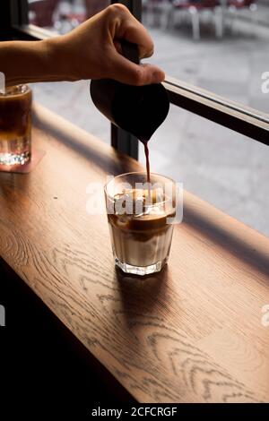 Ritagliare il cameriere versando il caffè fresco in un bicchiere con latte ghiacciato sul banco di legno vicino alla finestra nel cafe' Foto Stock