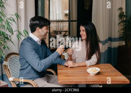 Felice giovane romantica affettuosa coppia tostatura con bicchieri di vino e tenendo le mani mentre si siede al tavolo di legno in accogliente caffè Foto Stock