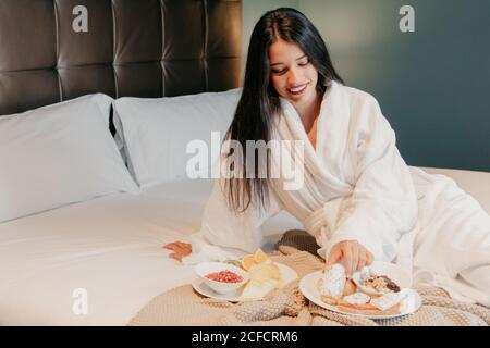 Felice giovane donna rilassata in accappatoio bianco seduto sul letto e gustando una deliziosa colazione in camera Foto Stock