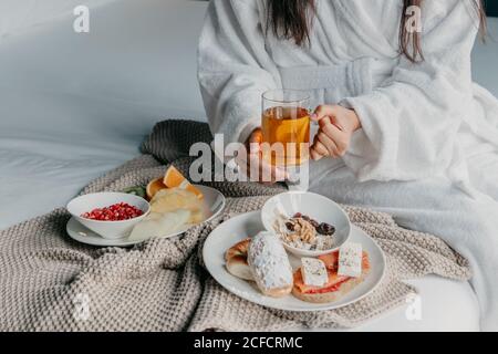 Brunette femmina tagliata e irriconoscibile in accappatoio bianco con tazza di vetro di tè mentre facendo colazione a letto con frutta e. pasticceria Foto Stock