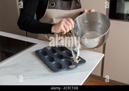 Coltivare la femmina nel grembiule tenendo la ciotola in acciaio inossidabile e il cucchiaio e. Versando l'impasto su una speciale muffa da forno per i biscotti Madeleine preparare il dessert al tavolo in marmo in cucina Foto Stock