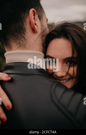 Giovane donna innamorata abbracciando un ragazzo irriconoscibile e sorridendo mentre guardava la macchina fotografica durante il giorno Foto Stock