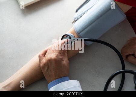 Dall'alto del medico del raccolto con stetoscopio seduto al tavolo in sala medica e la misurazione della pressione sanguigna con tonometro mentre lavorare in ospedale durante l'epidemia di coronavirus Foto Stock