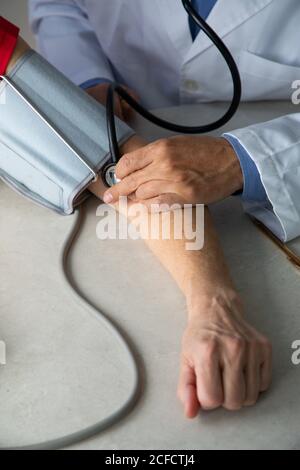 Dall'alto del medico del raccolto con stetoscopio seduto al tavolo in sala medica e la misurazione della pressione sanguigna con tonometro mentre lavorare in ospedale durante l'epidemia di coronavirus Foto Stock