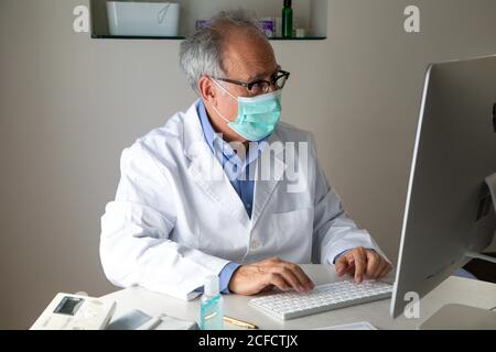 Medico di sesso maschile senior che indossa maschera facciale e abito medico seduto al tavolo e parlare con il paziente tramite videochiamata attiva Computer mentre lavora in remoto durante lo scoppio di COVID 19 Foto Stock