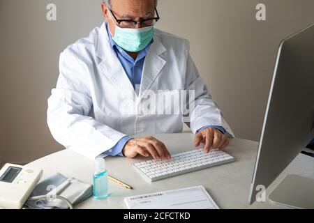 Medico di sesso maschile senior che indossa maschera facciale e abito medico seduto al tavolo e parlare con il paziente tramite videochiamata attiva Computer mentre lavora in remoto durante lo scoppio di COVID 19 Foto Stock