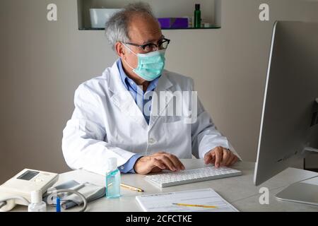 Medico di sesso maschile senior che indossa maschera facciale e abito medico seduto al tavolo e parlare con il paziente tramite videochiamata attiva Computer mentre lavora in remoto durante lo scoppio di COVID 19 Foto Stock