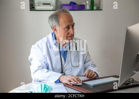 Medico maschile senior che indossa abito medico seduto a tavola e. parlare con il paziente tramite videochiamata sul computer mentre si lavora In remoto durante l'epidemia di COVID 19 Foto Stock