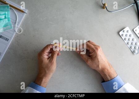 Vista dall'alto delle mani di un medico anziano seduto al tavolo in clinica e controllare il termometro mentre si lavora in ospedale durante pandemia di coronavirus Foto Stock