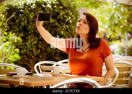 Donna adulta in abbigliamento casual che porta un selfie con il telefono e fare faccia a kissy mentre si siede su un accogliente caffè estivo terrazza Foto Stock