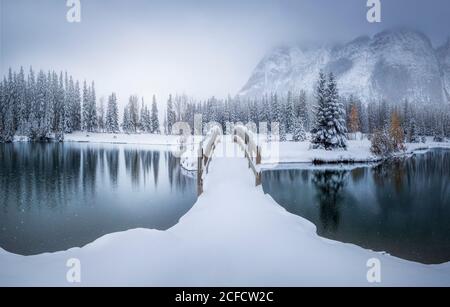 Splendido paesaggio invernale canadese con ponte innevato sulla calma acqua con foresta di abeti e nebbie montagne innevate sullo sfondo Foto Stock