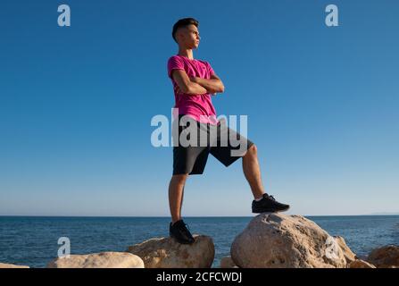 Da sotto di calmo maschio etnico con braccia incrociate in piedi sulla pietra vicino al mare contro il cielo blu chiaro che guarda via Foto Stock