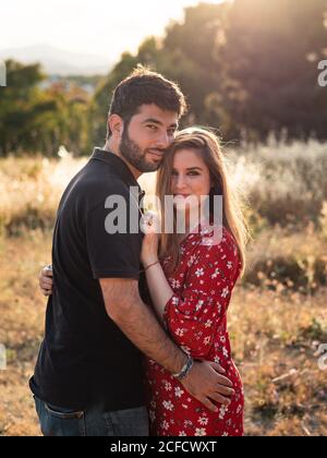 uomo che abbraccia teneramente sorridente moglie incinta sullo sfondo di pittoresco parco verde in giorno di sole Foto Stock