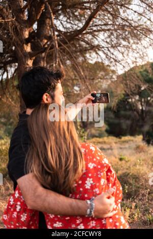 Felice giovane coppia baciando e prendendo selfie durante la camminata con un piccolo cane nel parco estivo Foto Stock