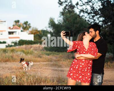 Felice giovane coppia baciando e prendendo selfie durante la camminata con un piccolo cane nel parco estivo Foto Stock