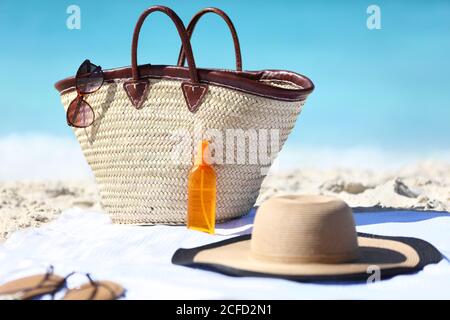 Accessori da spiaggia da donna sulla sabbia per un concetto di vacanza estiva. Borsa di paglia, cappello e crema solare o olio abbronzante spray bottiglia con Foto Stock