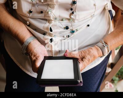 Dall'alto della donna anziana dai capelli grigi in camicia e bicchieri seduti su poltrona e leggendo un e-book in appartamento Foto Stock