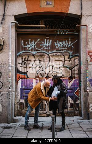 uomo adulto inginocchiato e baciante pancia di donna incinta contro gruggioso muro di graffiti sulla strada della città Foto Stock