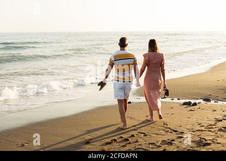 Vista posteriore di uomo e donna a piedi nudi che tiene le mani e che trasporta le scarpe mentre cammina sulla spiaggia sabbiosa verso il mare ondeggiante sul resort Foto Stock