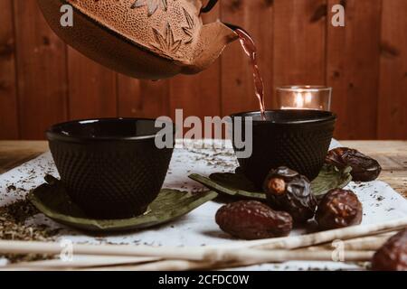 Serve tè saporito e fragrante in tazza di teiera in argilla e dolce datteri su vassoio bianco decorato con foglie di tè su legno sfondo Foto Stock