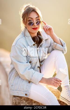 Donna con capelli biondi soddisfatta in occhiali da sole alla moda e abbigliamento casual seduto su una recinzione rocciosa e guardando la fotocamera alla luce del sole su sfondo sfocato Foto Stock