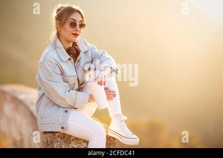 Donna con capelli biondi soddisfatta in occhiali da sole alla moda e abbigliamento casual seduto su una recinzione rocciosa e guardando la fotocamera alla luce del sole su sfondo sfocato Foto Stock