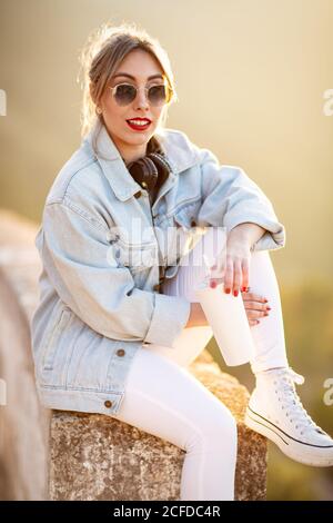 Donna con capelli biondi soddisfatta in occhiali da sole alla moda e abbigliamento casual seduto su una recinzione rocciosa e guardando la fotocamera alla luce del sole su sfondo sfocato Foto Stock