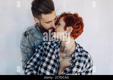 Positivo giovane coppia hipster con tatuaggi godendo il tempo insieme e. abbracciando mentre si levano in piedi contro la costruzione della pietra di shabby in giorno soleggiato Foto Stock