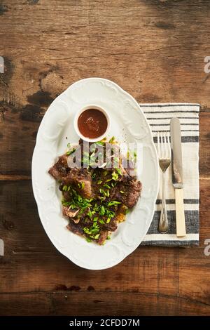 Vista dall'alto della gustosa carne al forno con cipolla verde E salsa barbecue su piatto con posate su legno rustico tabella Foto Stock