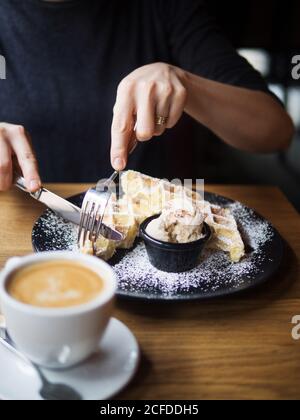 persona irriconoscibile che usa forma e coltello per tagliare i cialde dolci vicino alla ciotola del gelato e a una tazza di caffè tavolo in caffetteria Foto Stock
