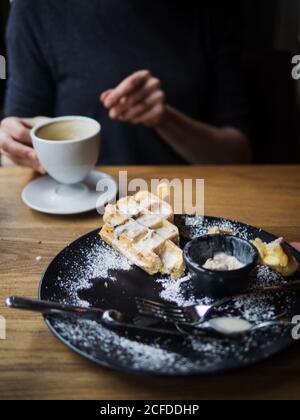 Piatto con resti di waffle e gelato tavolo da caffetteria vicino a una persona irriconoscibile che beve caffè Foto Stock