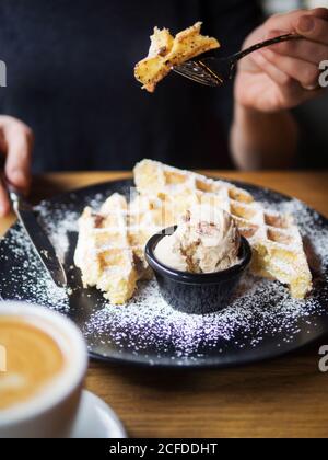 persona irriconoscibile che usa forma e coltello per tagliare i cialde dolci vicino alla ciotola del gelato e a una tazza di caffè tavolo in caffetteria Foto Stock