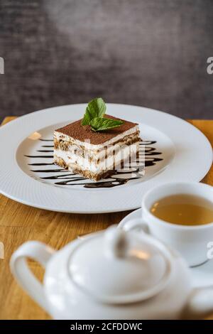 Gustoso e appetitoso tiramisù al cioccolato decorato con verde e versato con sciroppo su piatto bianco e tè in tazza bianca con teiera sul tavolo nel ristorante Foto Stock