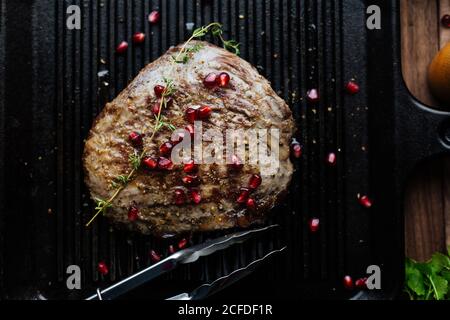 Vista dall'alto della bistecca di manzo appetitosa guarnita con semi di melograno ed erbe servite su piastra nera con le pinze della cucina sopra tavolo di legno Foto Stock