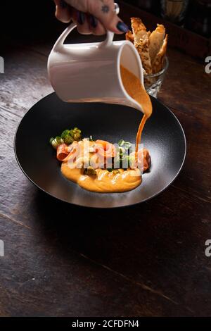 Dall'alto della mano di femmina chef che versa piatto con pesce rosso ed erbe verdi con salsa d'arancia in elegante ciotola nera Foto Stock