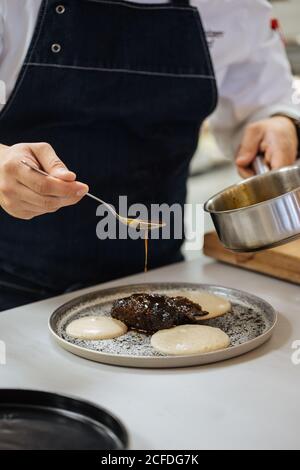 Crop chef ristorante con pentola e cucchiaio in mani versando condite con il cibo mentre preparate un elegante piatto di alta cucina Foto Stock