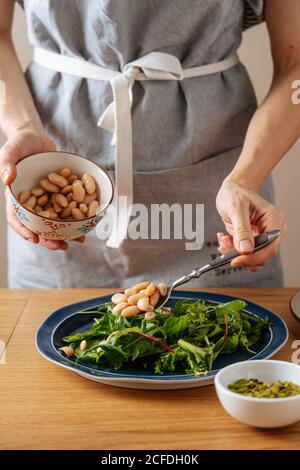 Crop Woman in grembiule mettere fagioli bianchi su erbe fresche mentre preparare insalata vegetariana sana per il pranzo Foto Stock