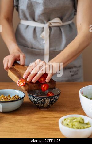 donna irriconoscibile in grembiule che mette i pomodori ciliegini tagliati in ciotola durante la preparazione dell'insalata vegana sul tavolo in cucina Foto Stock