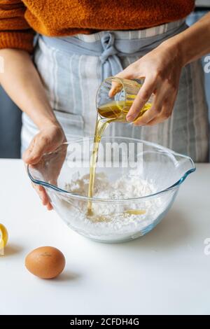 Coltivare l'olio d'oliva femminile che fuoriesce dal vetro in un recipiente grande con farina mentre si prepara l'impasto al tavolo bianco in cucina Foto Stock