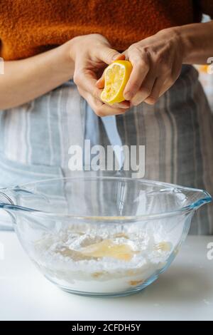 Crop femmina casual in grembiule spremitura limone su vetro trasparente ciotola con ingredienti misti mentre prepari l'impasto in cucina Foto Stock