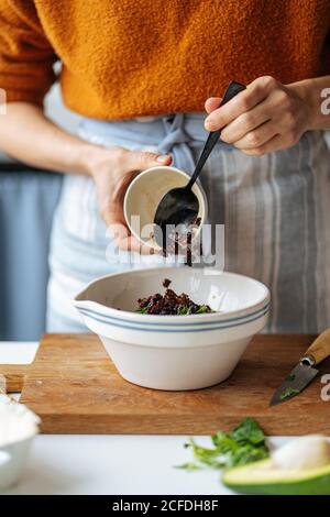 Crop femmina aggiungendo uvetta nera in ceramica bianca recipiente posto su tagliere di legno mentre preparano il cibo alla cucina domestica Foto Stock