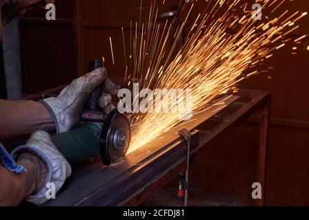 Mani tagliate di un uomo da lavoro in occhiali protettivi e guanti da taglio metallo con smerigliatrice con scintille volanti durante il lavoro in officina Foto Stock