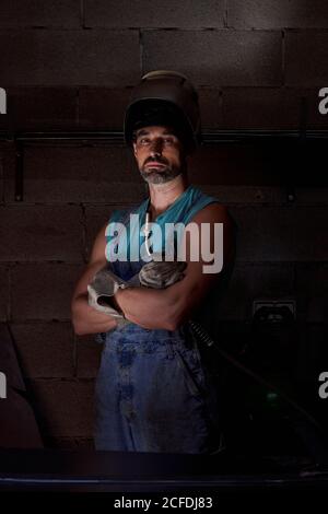 Uomo di lavoro con la bearded serio sicuro nel casco di saldatura sulla testa che indossa tute e guanti di protezione in piedi con braccia incrociate al buio muro di pietra sullo sfondo Foto Stock