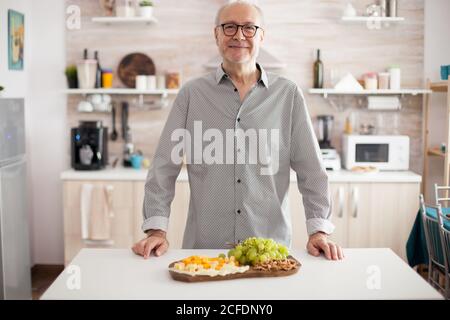 Allegro uomo maturo in cucina sorridente a macchina fotografica con formaggi assortiti e uva. Foto Stock